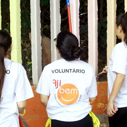 Três voluntárias estão olhando para cima, de costas, com camisetas AJIBEM